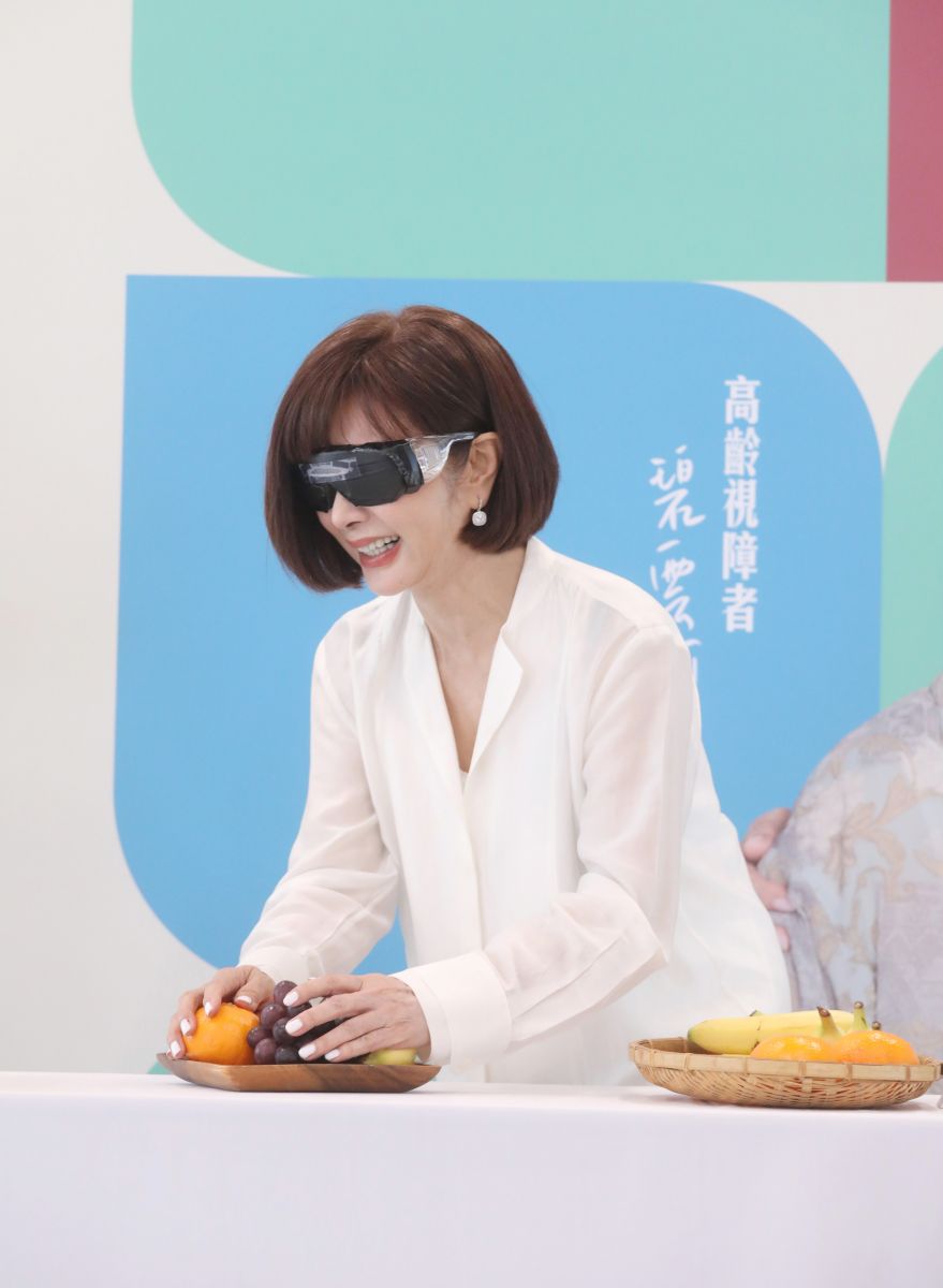 愛盲基金會年度代言人陳美鳳在記者會上戴上視覺模擬眼鏡進行闖關,找出水果擺盤和日用品